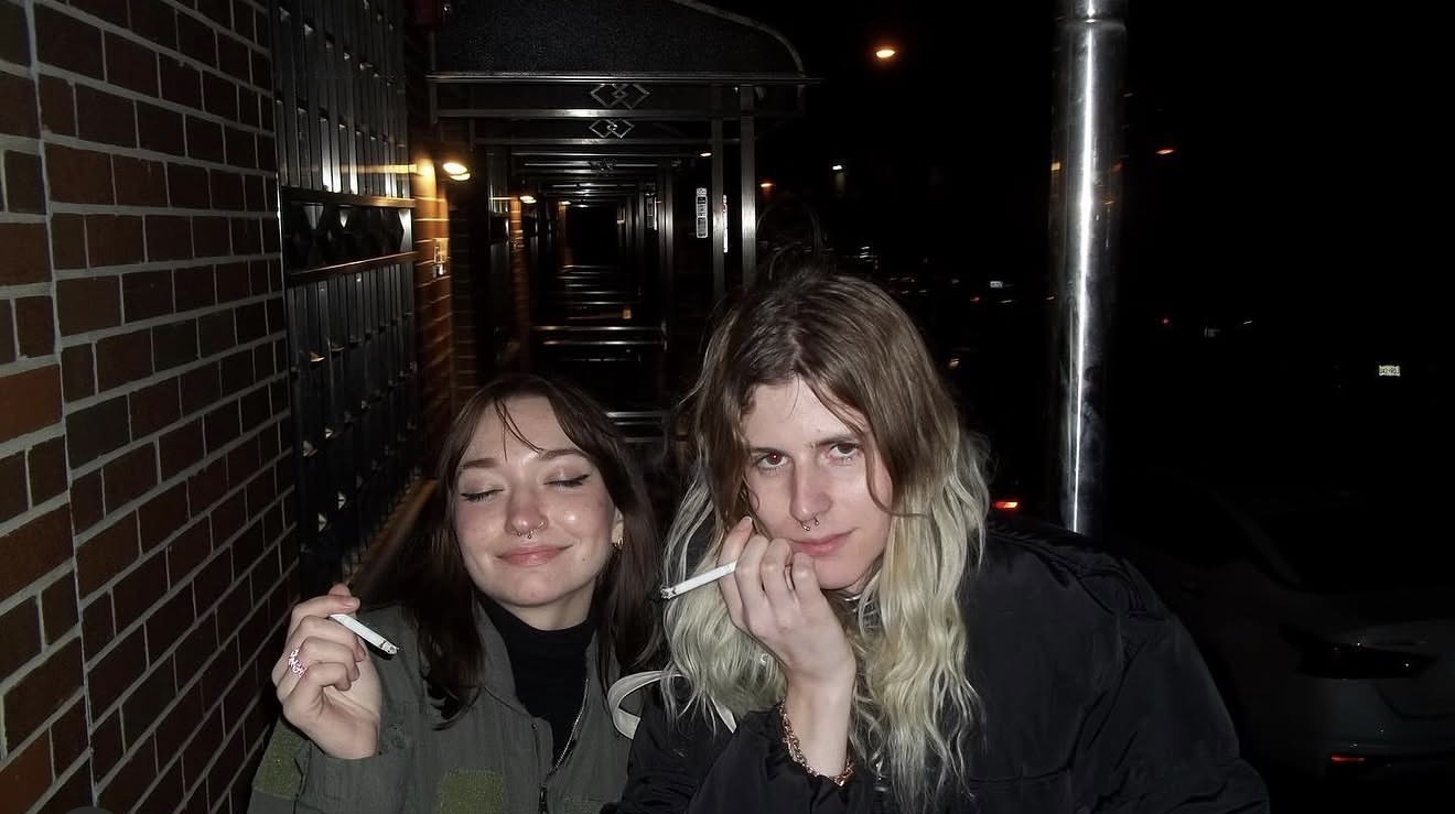 Anastasiya with a friend outside at night, sharing a cigarette on a brick-lined sidewalk